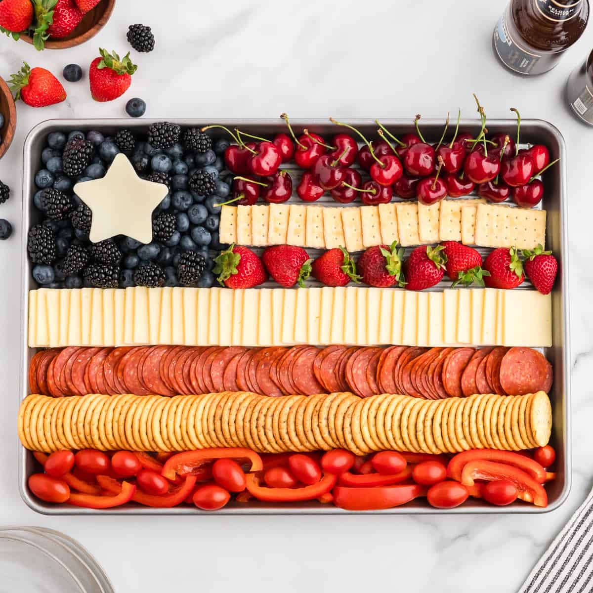 A sheet pan with a charcuterie board in the shape of the American flag. The flag has alternating red and white strips of cheese, crackers, pepperoni, fruits and vegetables. The top right is a blue section of blueberries and blackberries with a white cheese star.