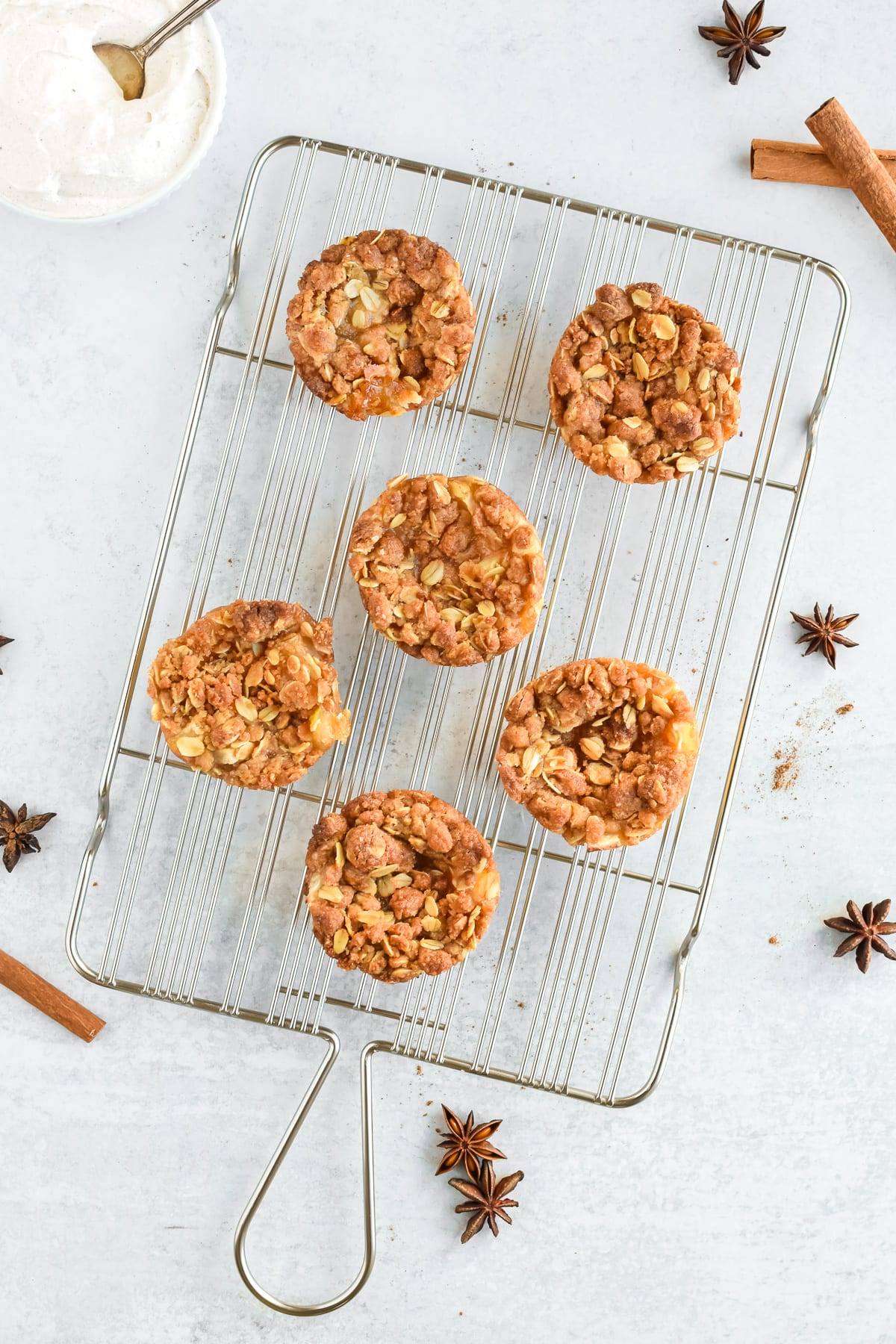 A cooling rack with 6 mini apple crumble tarts. Cinnamon sticks and star anise are to the side, along with a bowl of whipped cream.