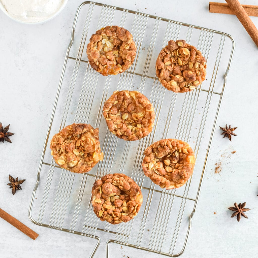 A cooling rack with 6 mini apple crumble tarts. Cinnamon sticks and star anise are to the side, along with a bowl of whipped cream.