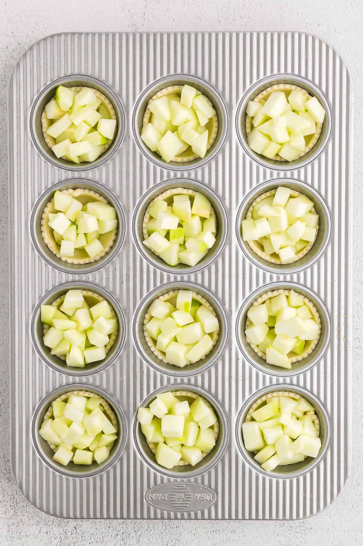 A muffin pan with pie crust and diced apples in each cup.