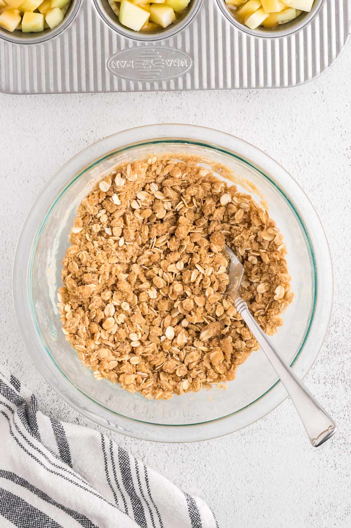 A glass bowl with crumble topping in it, being stirred with a fork.