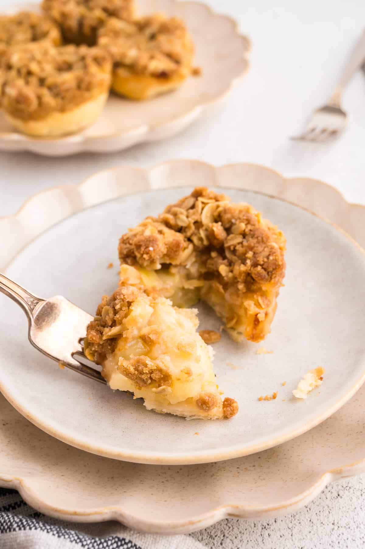 A close up of a fork with a slice of apple crumble tartlet on it, with more desserts in the background.