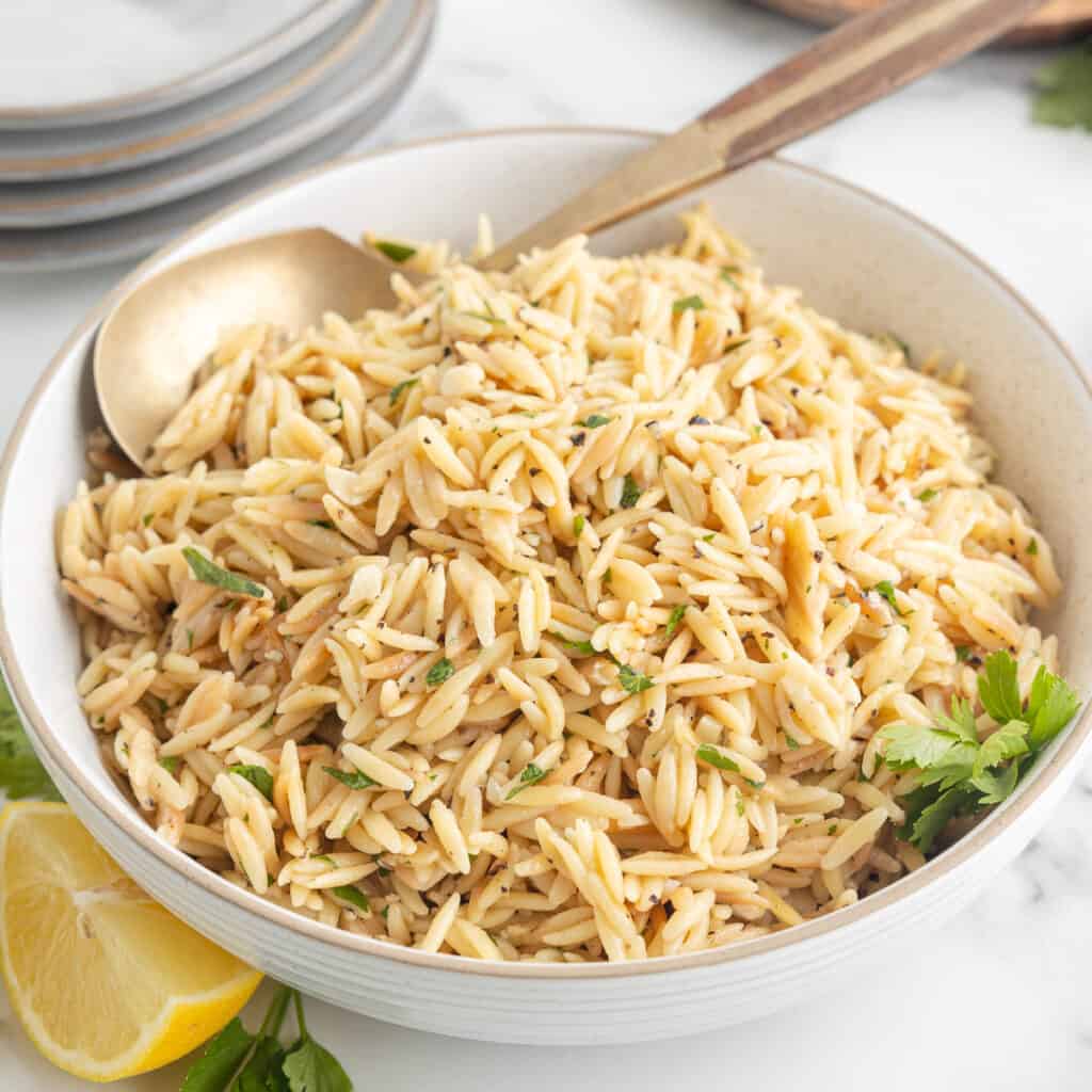 A close up side view of a serving dish of lemon garlic orzo. A spoon rests in the dish, and a quartered lemon and sprig of parsley are in the foreground.