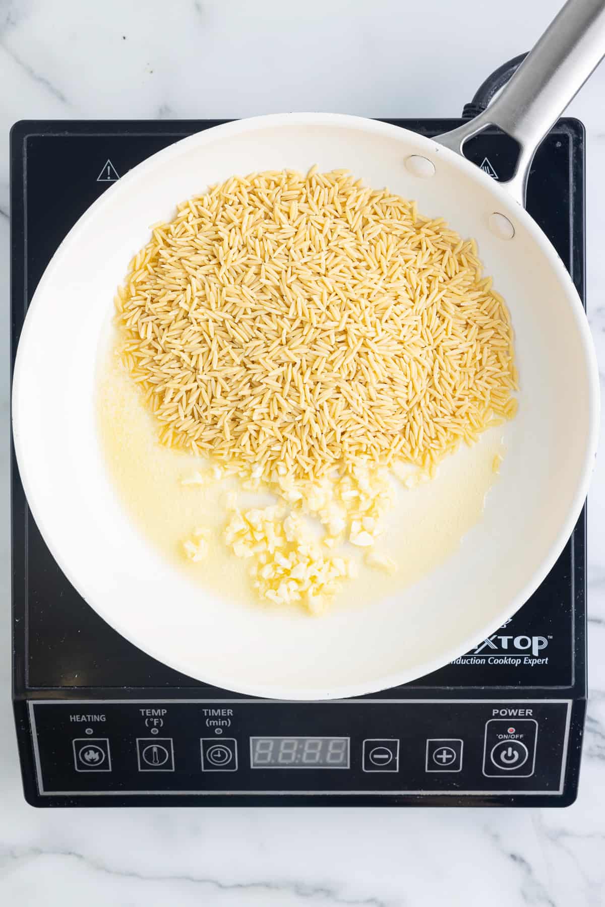 A white skillet on a black cooktop sitting on a marble background. The skillet has minced garlic and uncooked orzo pasta in it.