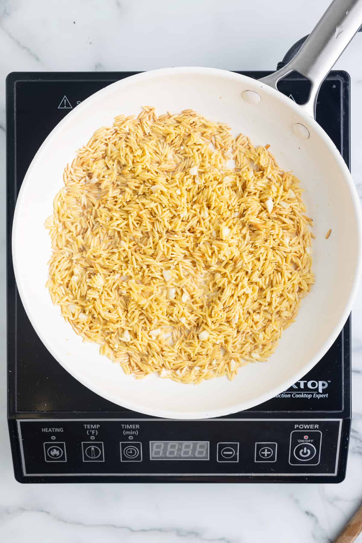 A white skillet on a black cooktop sitting on a marble background. The skillet has toasted orzo and minced garlic in it.