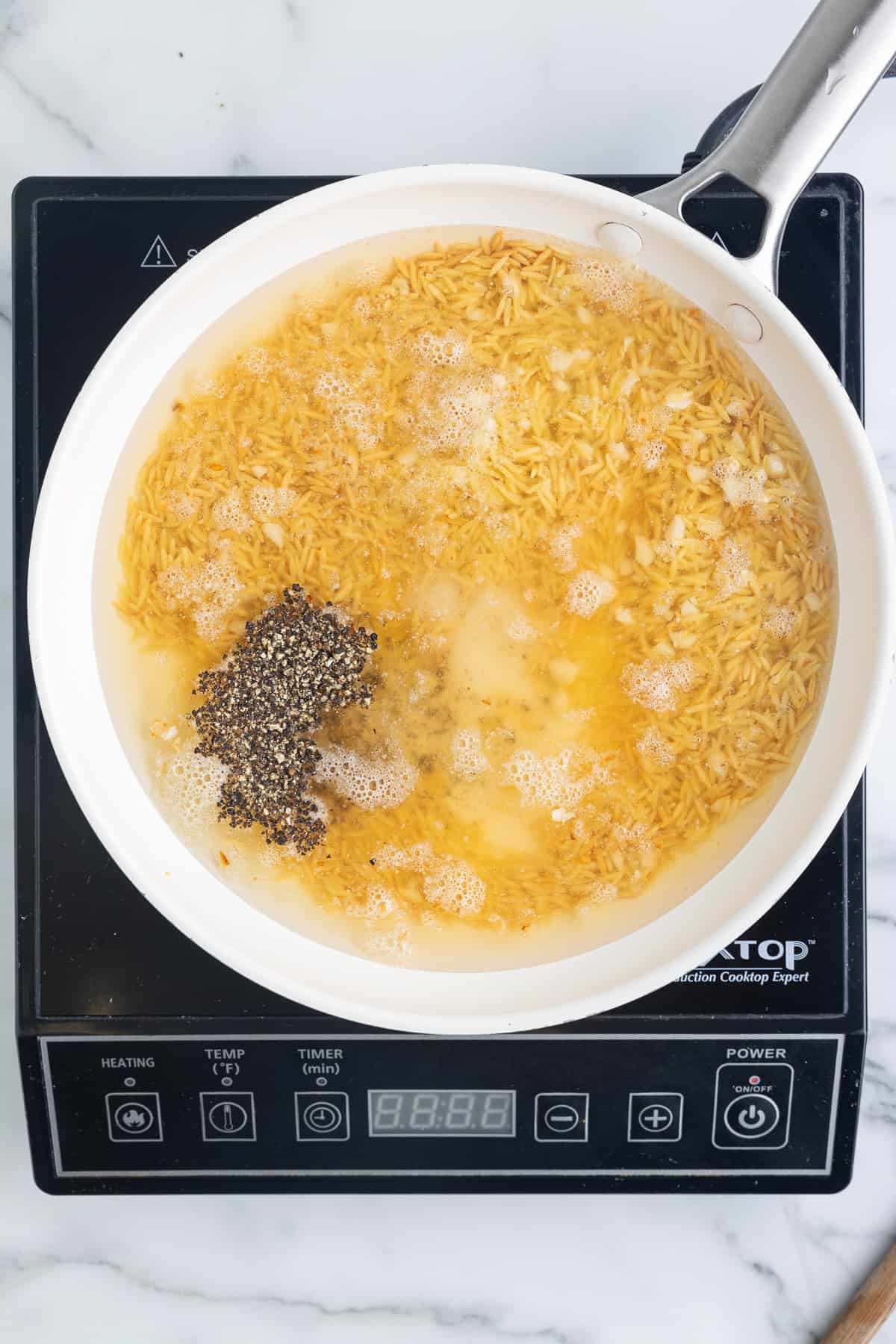 A white skillet on a black cooktop sitting on a marble background. The skillet has orzo covered in chicken stock, seasoned with salt and pepper.