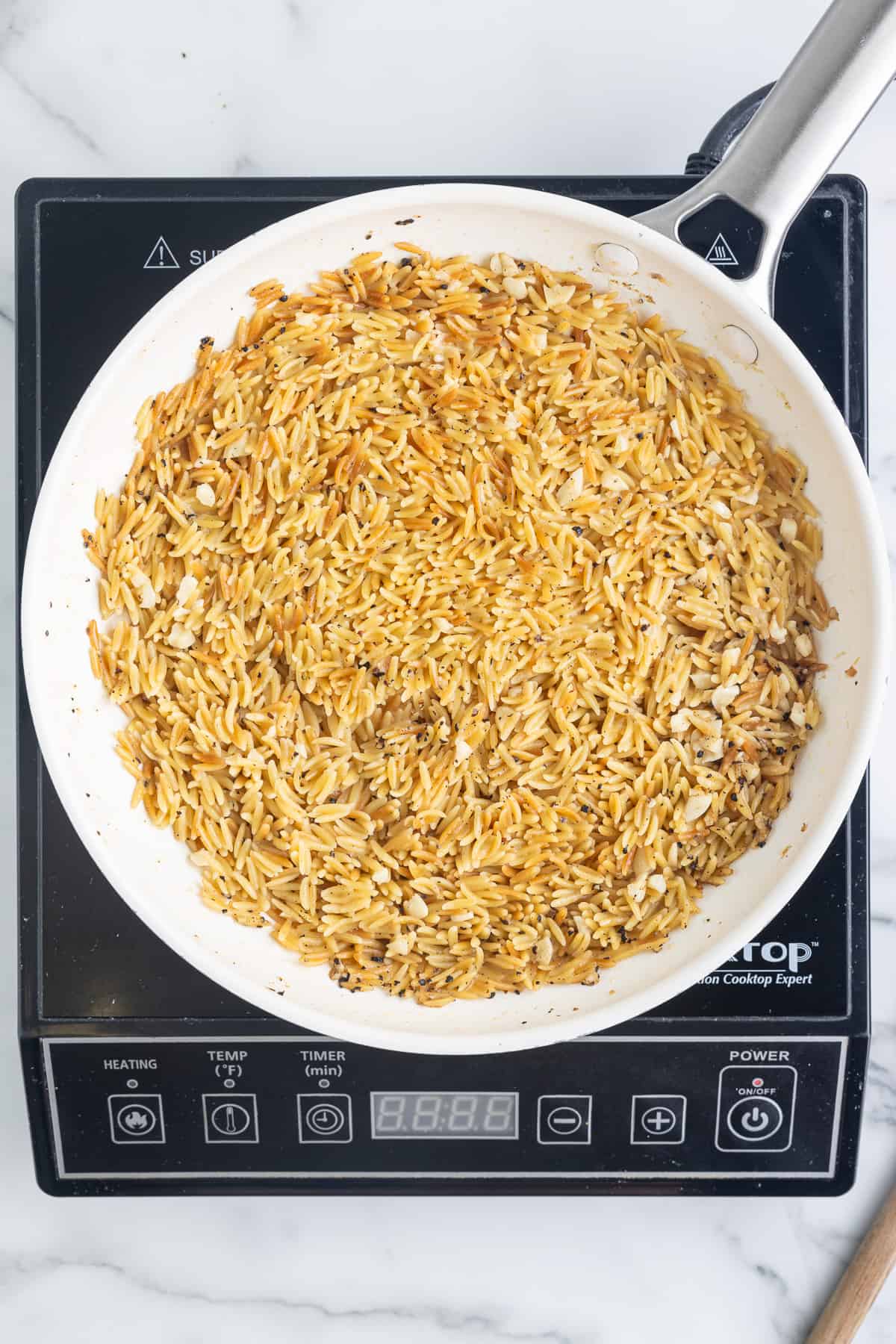 A white skillet on a black cooktop sitting on a marble background. The skillet has cooked garlic orzo in it.