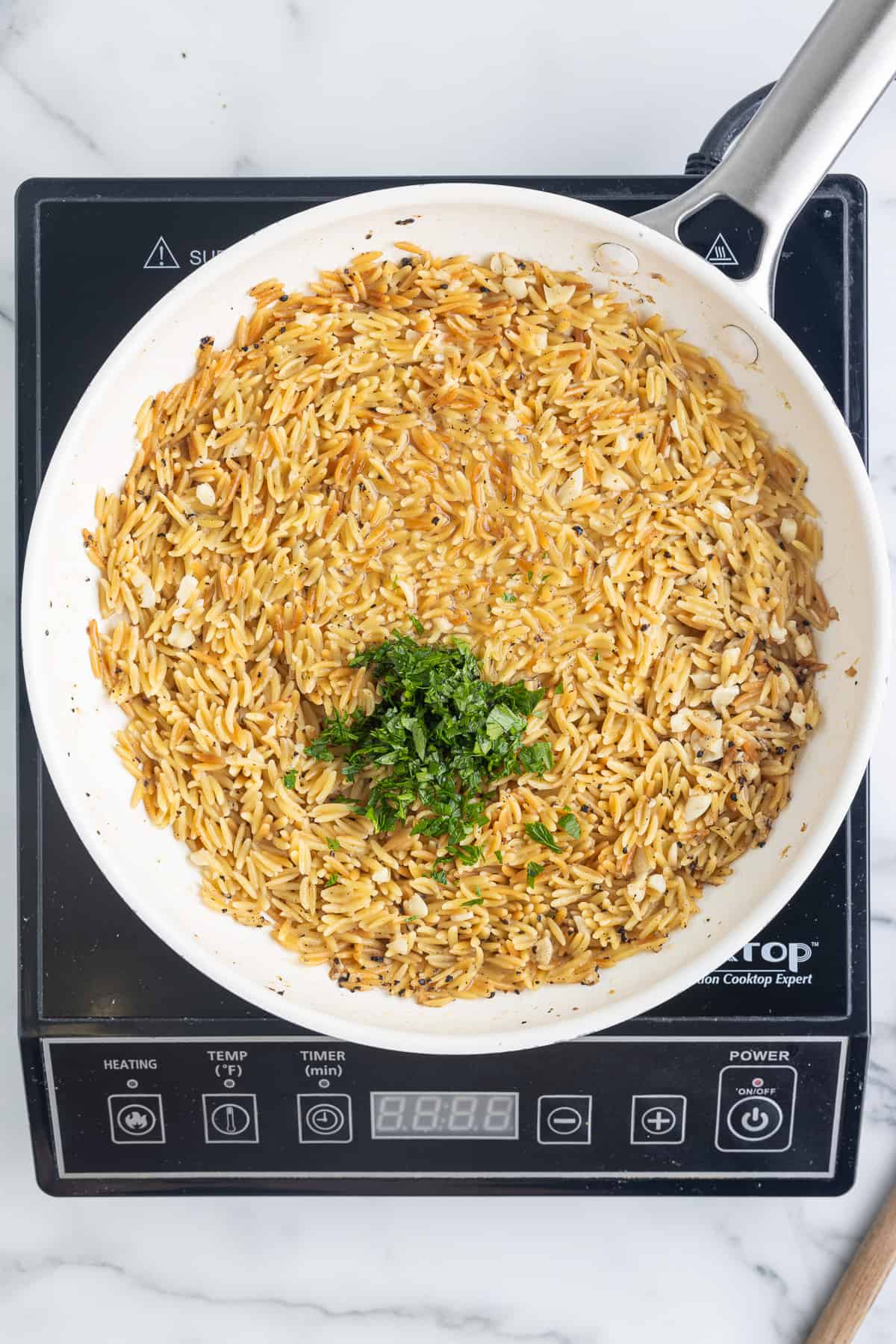 A white skillet on a black cooktop sitting on a marble background. The skillet has cooked orzo in it, topped with lemon juice and mined garlic.