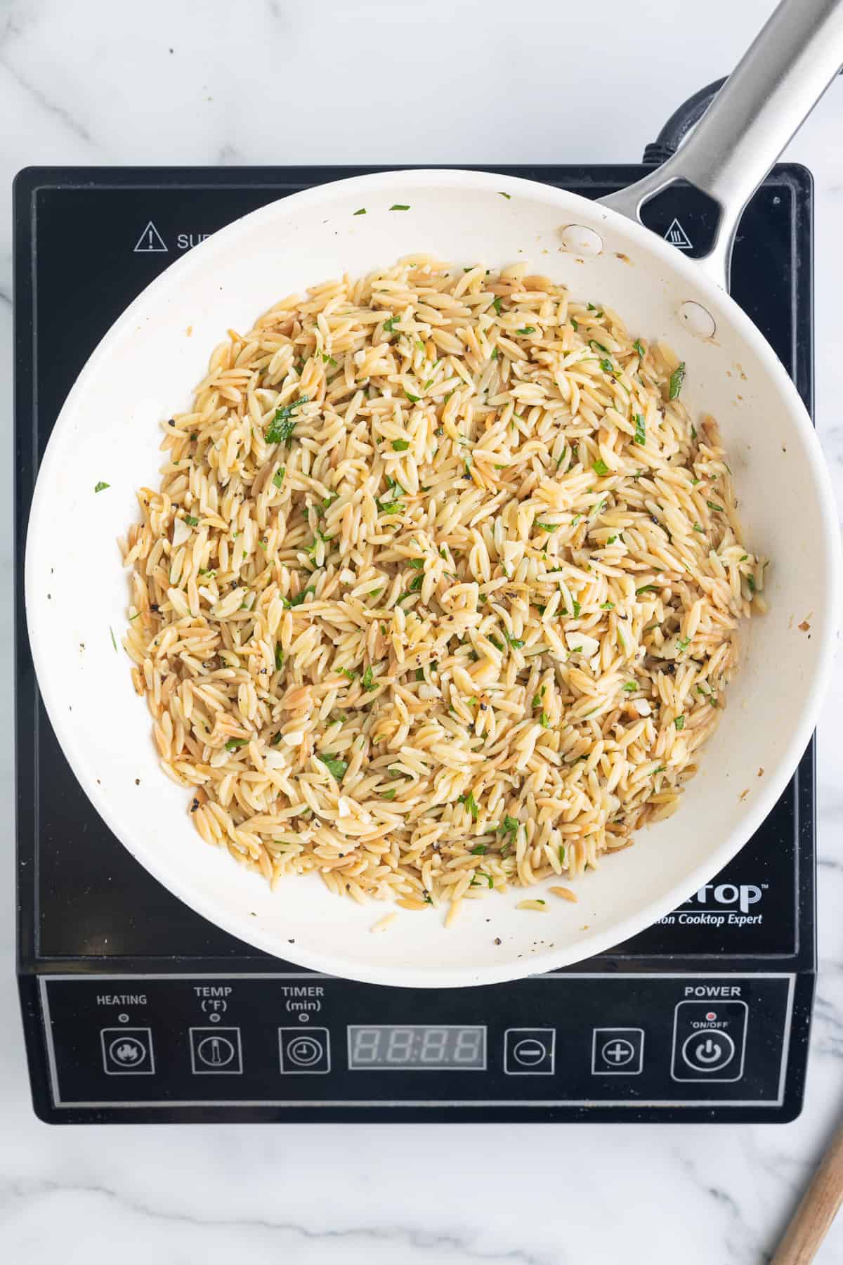 A white skillet on a black cooktop sitting on a marble background. The skillet has fully cooked lemon garlic orzo in it.