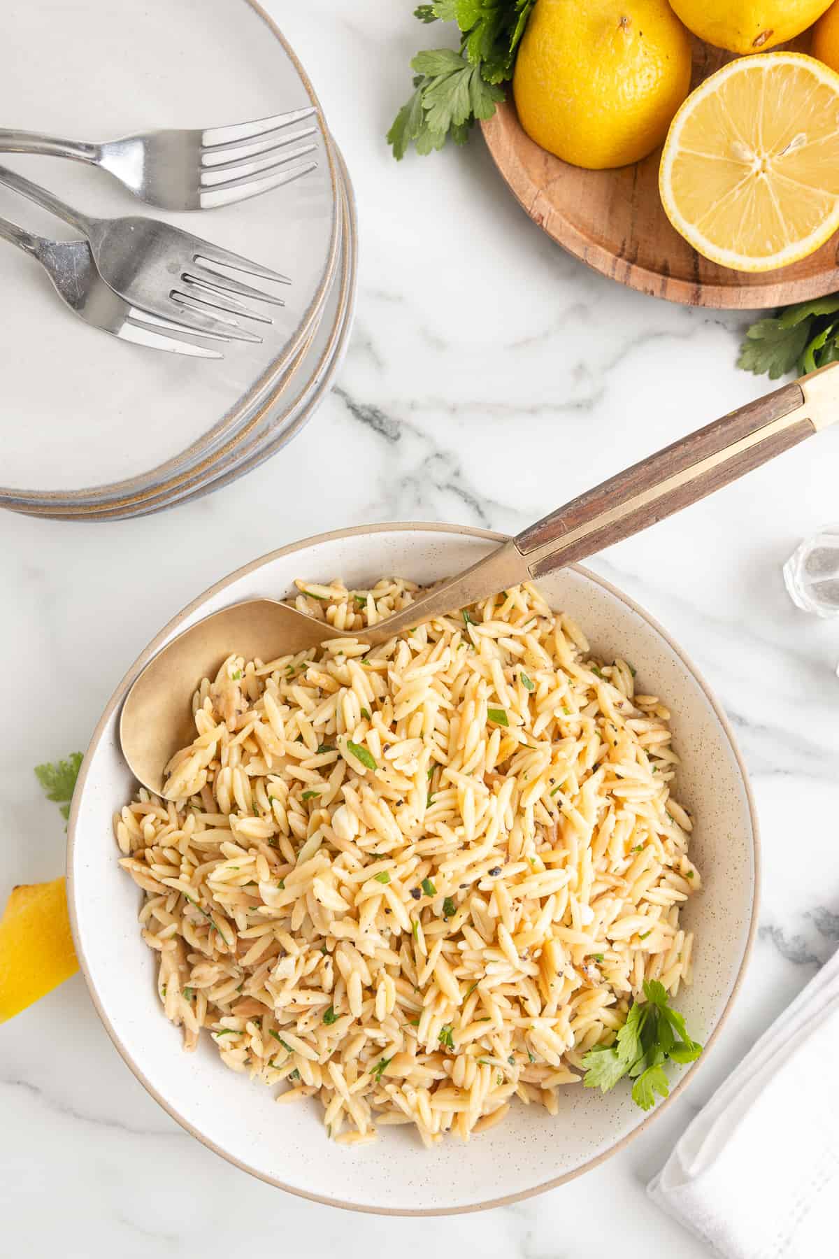 A white serving dish on a marble background. The dish contains cooked lemon garlic orzo. A wooden spoon rests in the dish, and three plates with forks and a dish of lemon and parsley sits to the side.