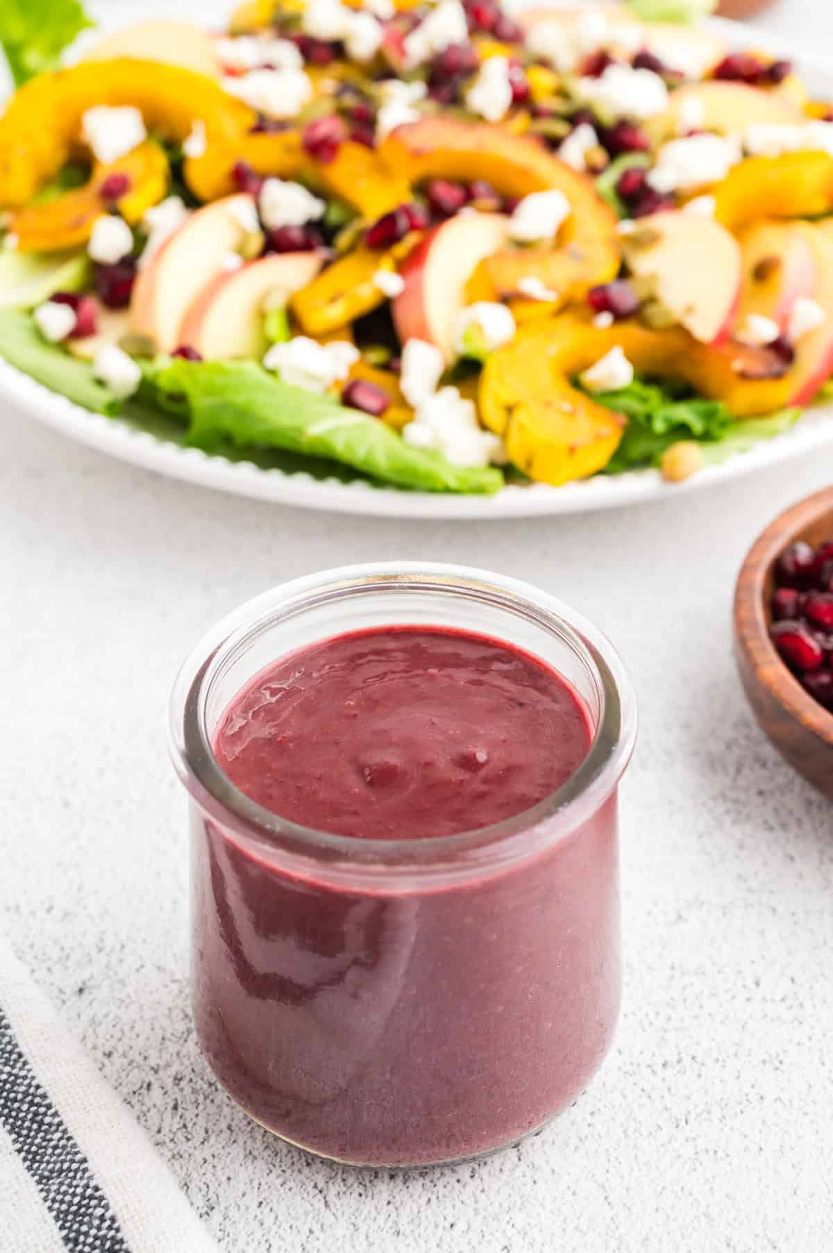 A jar of blackberry vinaigrette on a white concrete background. A colorful salad is in the background.