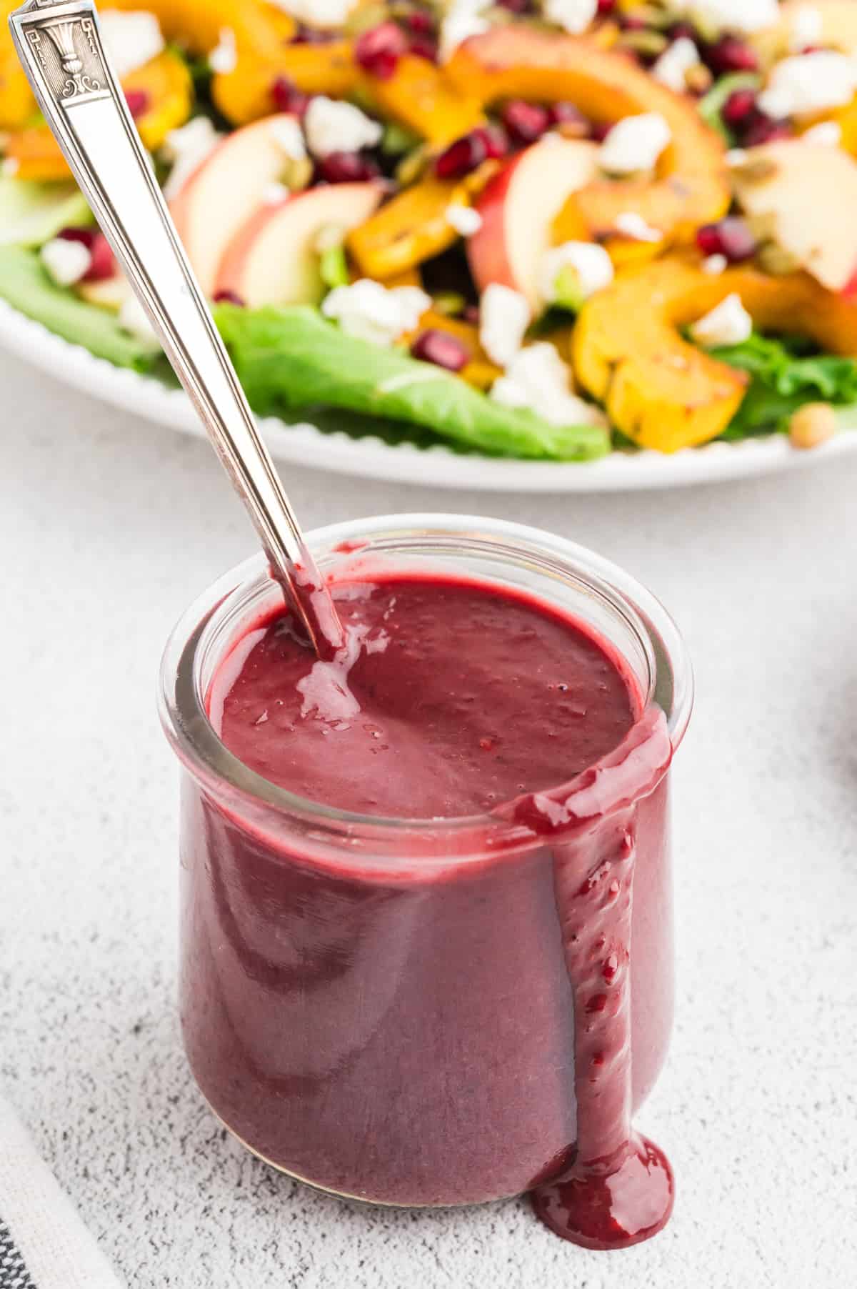 A jar of blackberry vinaigrette on a white concrete background. A drip of vinaigrette falls over the edge of the jar, and a spoon rests in it. A colorful salad is in the background.