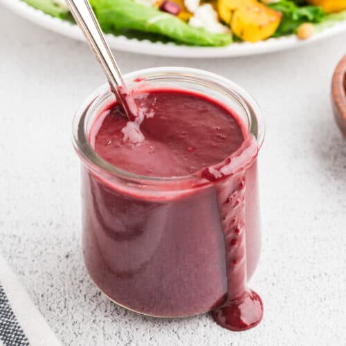 A jar of blackberry vinaigrette on a white concrete background. A drip of vinaigrette falls over the edge of the jar, and a spoon rests in it.