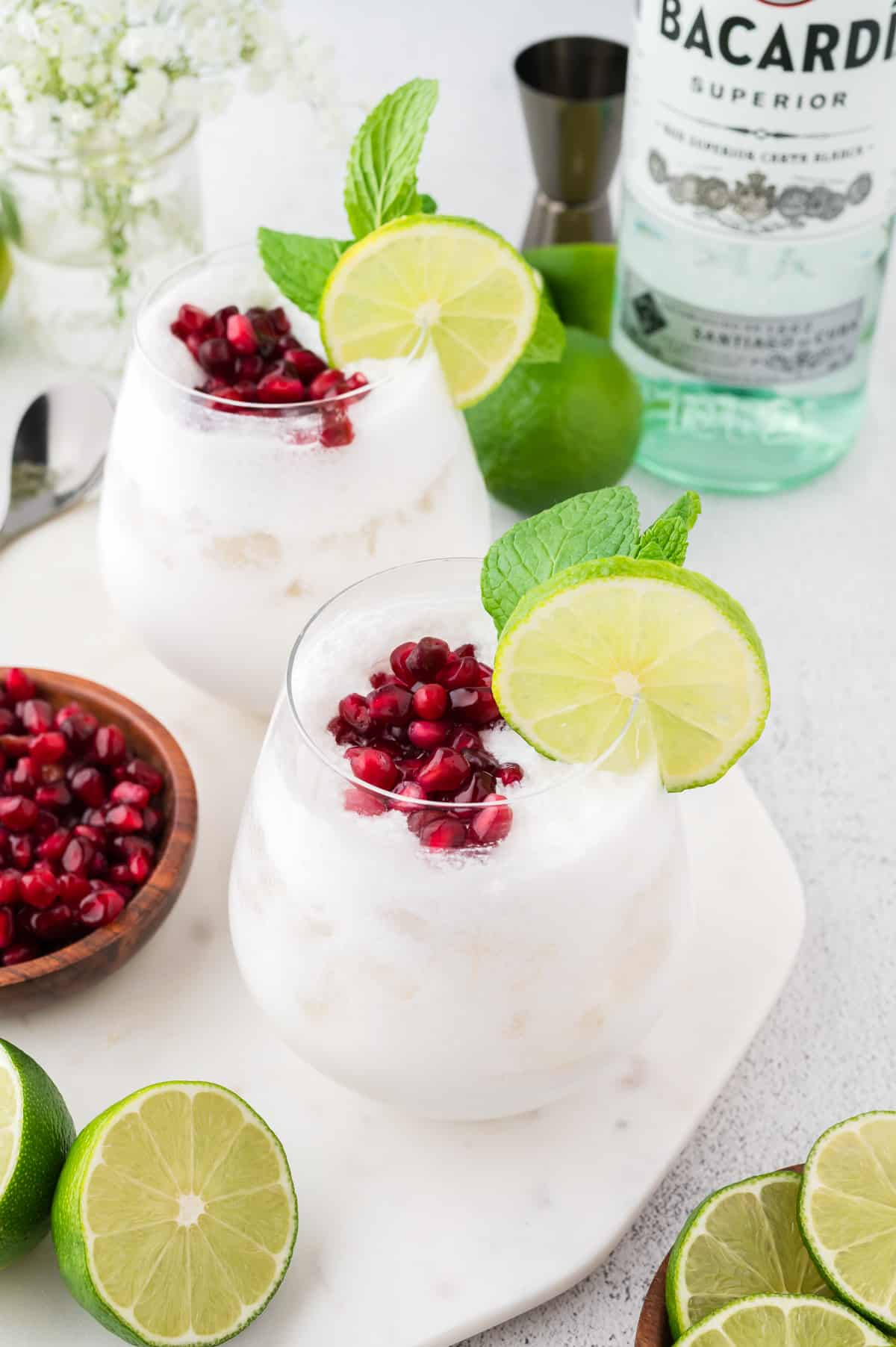 Two white coconut mojitos topped with pomegranate seeds and mint, with more pomegranate seeds, lime and mint to the side. A bottle of rum is in the background.