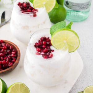 Two white coconut mojitos topped with pomegranate seeds and mint, with more pomegranate seeds, lime and mint to the side. A bottle of rum is in the background.