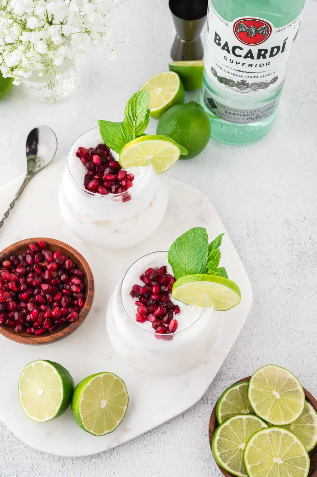 A top down view of two white coconut mojitos topped with pomegranate seeds and mint, with more pomegranate seeds, lime and mint to the side. A bottle of rum is in the background.