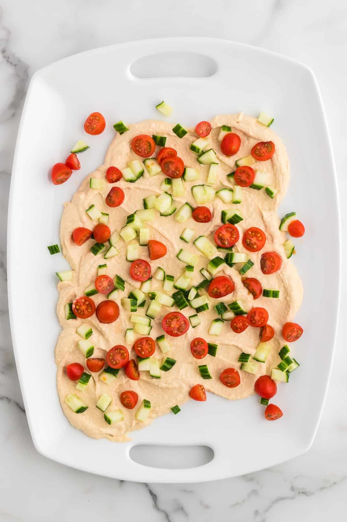 A white platter with hummus swirled on it, topped with cherry tomatoes and diced cucumbers.