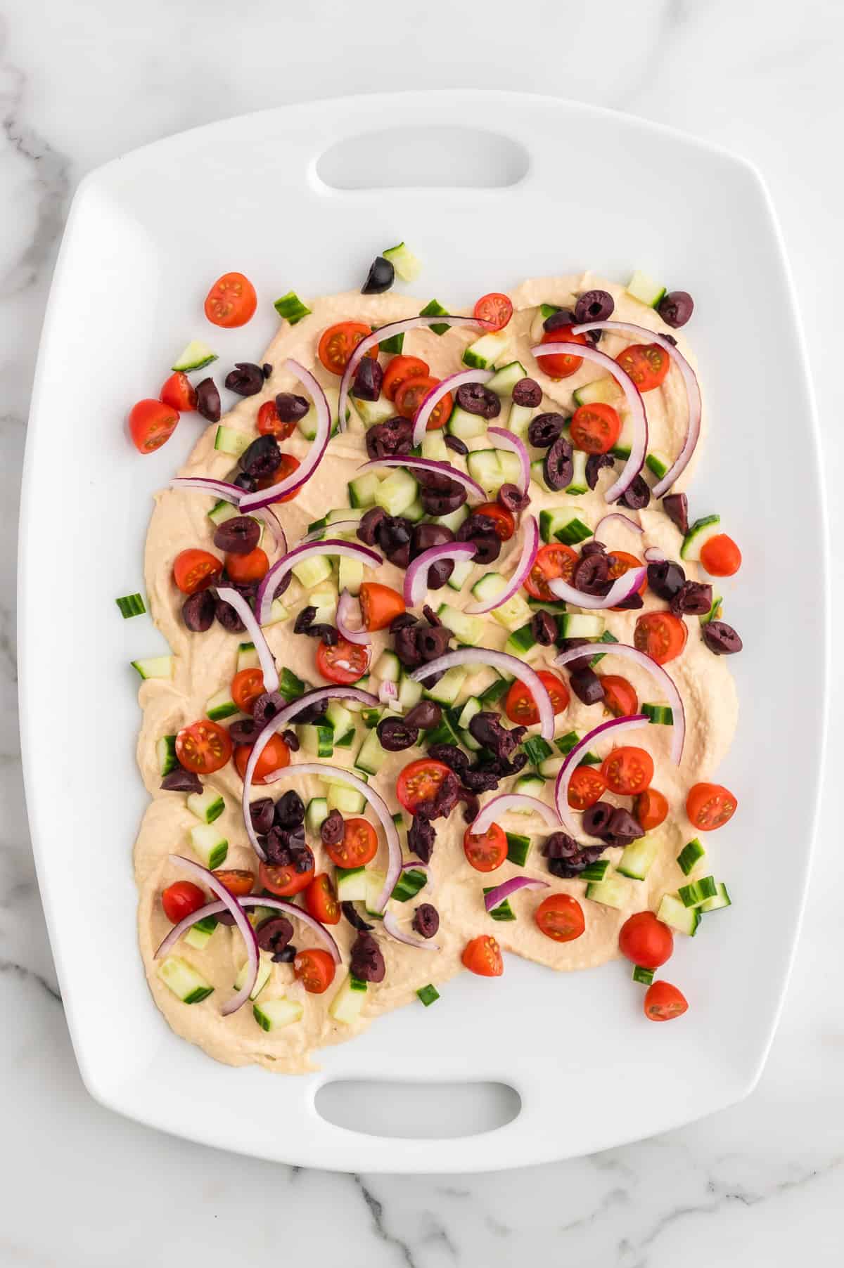 A white platter with hummus swirled on it, topped with cherry tomatoes, diced cucumbers, red onion and sliced kalamata olives.