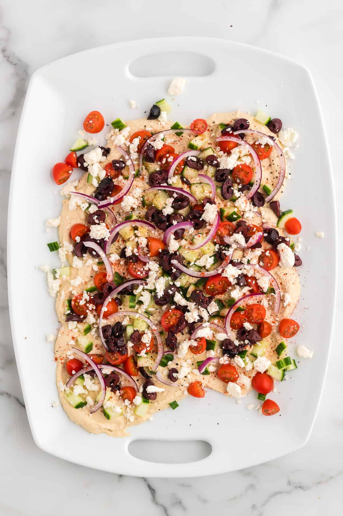 A white platter with hummus swirled on it, topped with cherry tomatoes, diced cucumbers, red onion, sliced kalamata olives, feta cheese and spices.