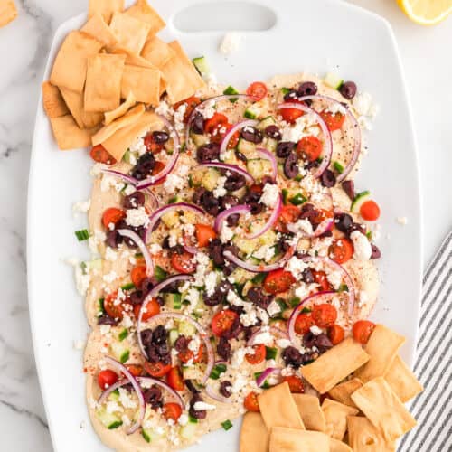 A white platter on a marble background with loaded hummus topped with cucumbers, cherry tomatoes, red onion, feta cheese and kalamata olives. Pita chips are arranged on the sides of the platter.